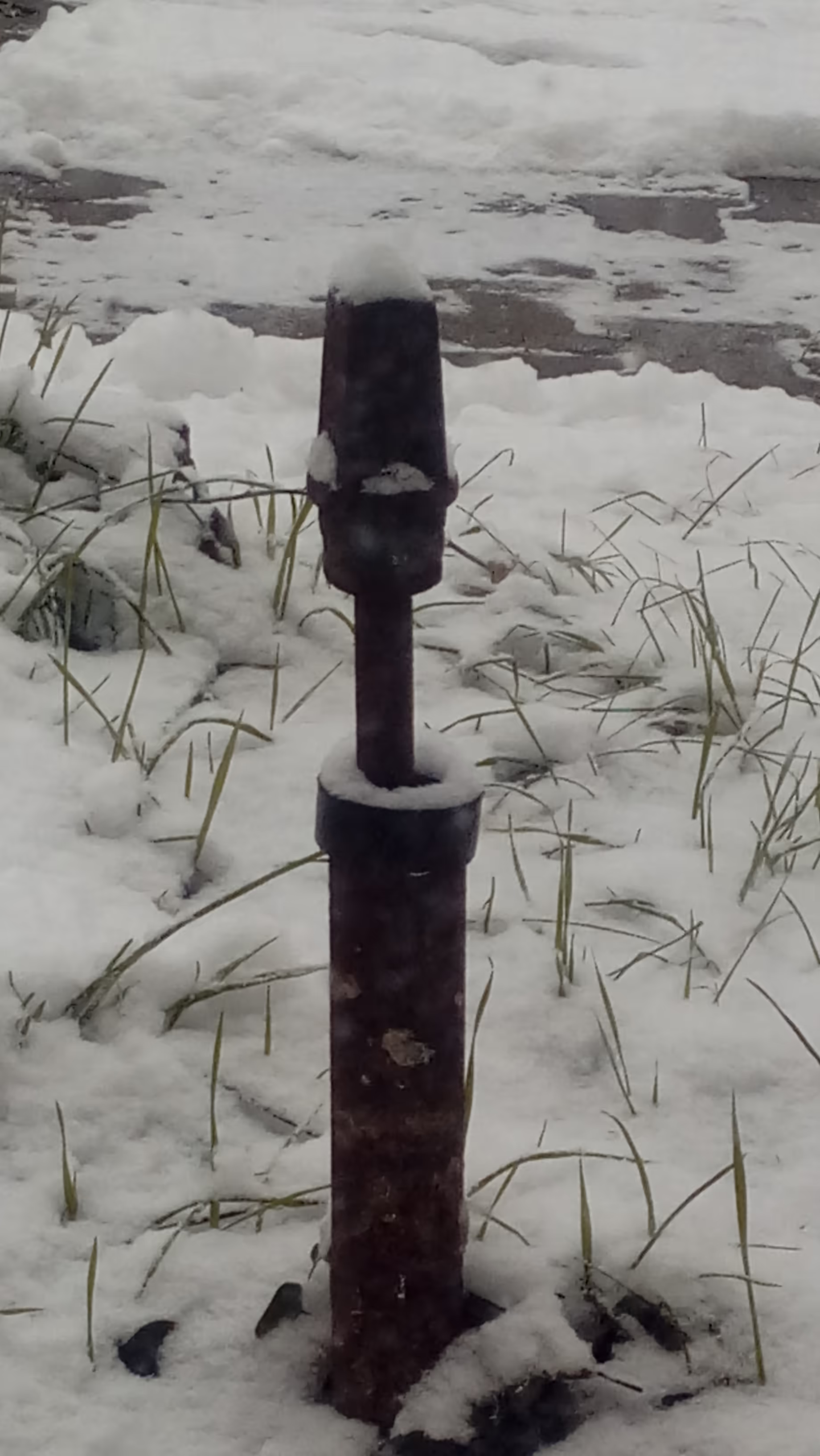 A rusty metal pipe sticking out of the ground. A smaller bar with a bigger metal piece on top is inside the pipe, also sticking up. Every surface of and the ground around the pipe is covered in snow. Next to the pipe some blades of grass stick out of the snow. In the background, a bit of pavement is visible because the snow has been shoveled.