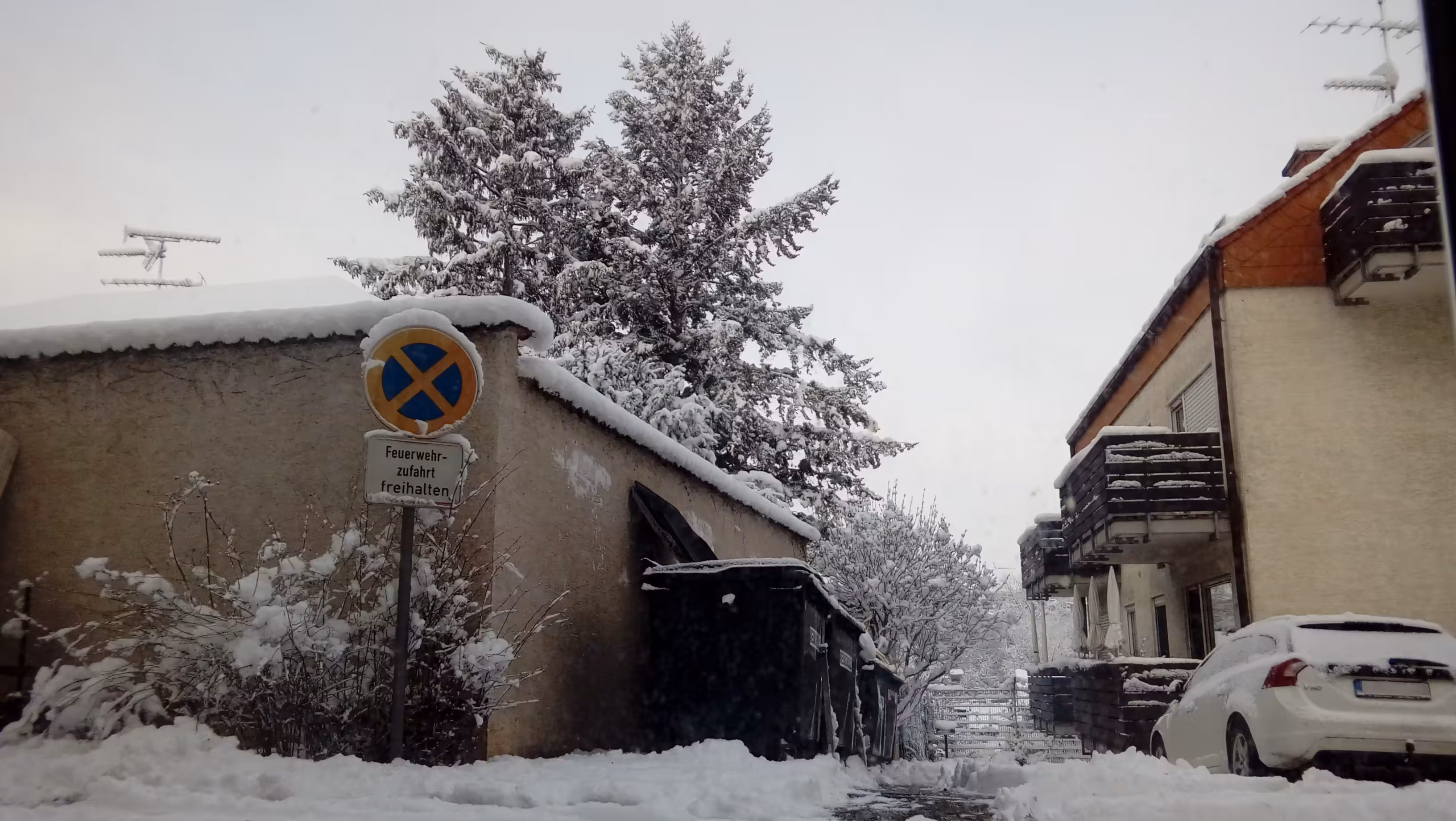 An outside scene where everything is covered in snow. On the left side, a grey-yellowish wall can be seen going around a corner. In front of it is a sign that sais, in german, "Feuerwehrzufahrt freihalten" and a few dead bushes with big clumps of snow between their twigs. To the right of the wall there are four big, black dumpsters, one of which is open. Behind the wall, the antanne of a house and two tall pinetrees stand against the grey sky, the dark branches of the latter contrasting with the snow that's piled up on them. On the right side of the picture, a house with balconies is visible. In front of it, a white car.