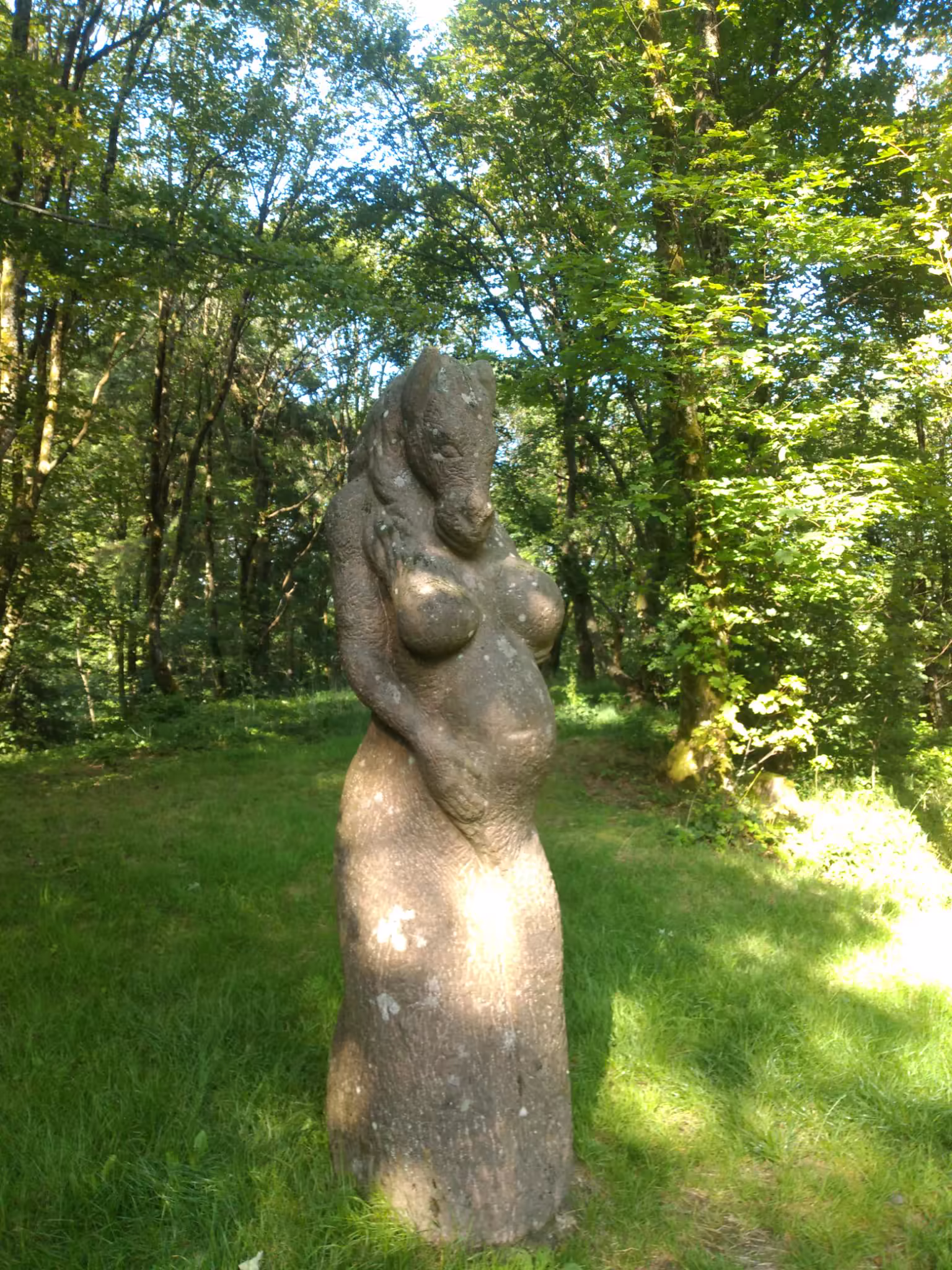 A rock statue of a pregnant woman with a horses head stands in the grass, sourrounded by trees. Her right arm can be seen holding her belly. The breasts on the statue are rather well-pronouned.